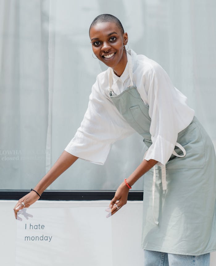 Happy female barista holding a sign with 'I hate Monday' outside a café. Cheerful and professional.