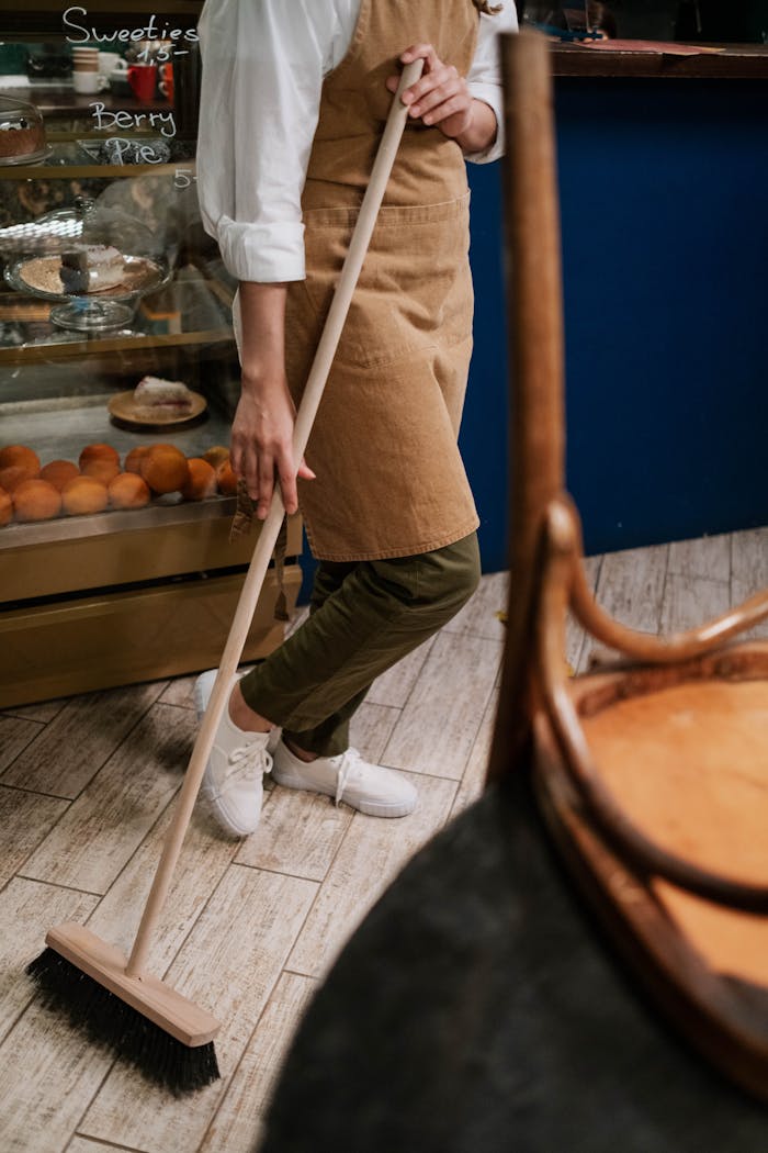 A person in an apron sweeps the wooden floor of a bakery with a broom, near pastries.